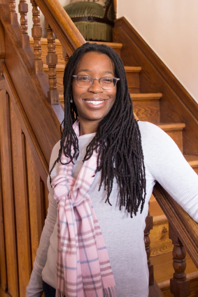 Tiya Miles smiling, wearing a pink scarf with her arm on historic wooden bannister with wood stairs in the background