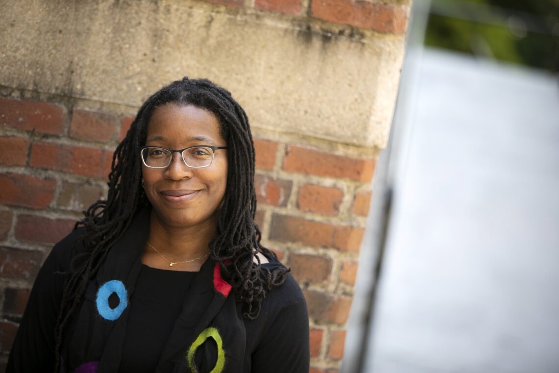 Tiya wearing a black sweater with blue, yellow, and red circles standing in front of a brick wall smiling