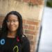 Tiya wearing a black sweater with blue, yellow, and red circles standing in front of a brick wall smiling